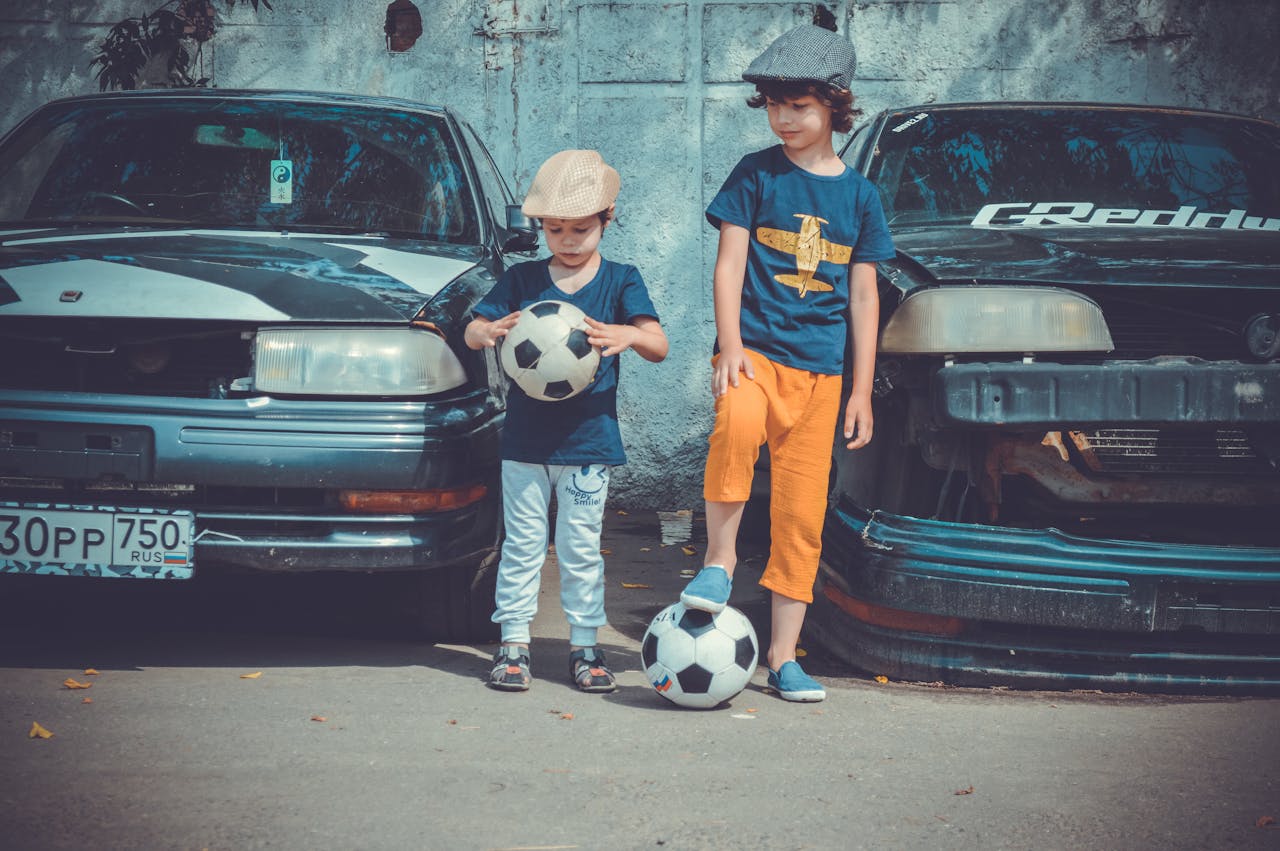 Two children in casual attire play soccer near vintage cars on a sunny day.